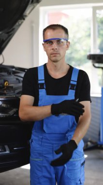 Vertical format video of young caucasian man in blue coveralls and protective glasses crosses arms, smiling and looking into camera. Male car mechanic at workplace in spacious repair shop.