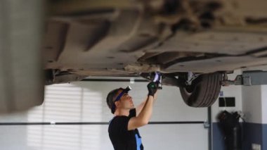 Auto mechanic working underneath car lifting machine at the garage. Auto repair shop. Male mechanic working on vehicle in car service. Man wearing gloves and using ratchet underneath the car.