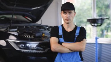 Portrait of a car mechanic crosses hands in a car workshop in blue uniform with equipment looking into camera.