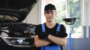 Young Caucasian man in blue coveralls, black cap and protective glasses crosses arms, smiling and looking into camera. Male car mechanic at workplace in spacious repair shop.