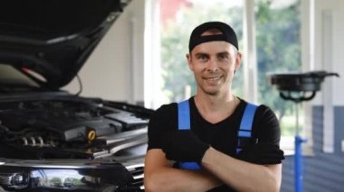 Portrait happy mechanic worker in blue uniform and cap from auto service center in front of the camera looking straight smiling and enjoying the time at his work place.