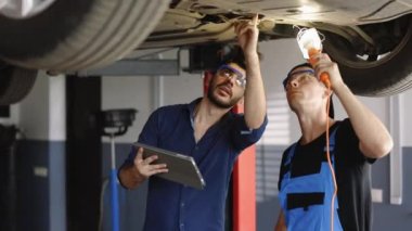 Manager Checks Data on a Tablet Computer and Explains the Breakdown to Mechanic. Car Service Employees Inspect the Bottom of the Car with a LED Lamp. Modern Workshop. Auto Service.