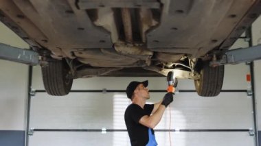 Mechanic in blue coveralls and black cap check out automobile parts while working with led lamp. Auto mechanic working underneath car lifting machine at the garage. Car auto service.