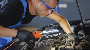 Young caucasian man in blue overalls and safety glasses inspects engine with flashlight. Male car mechanic at work in spacious repair shop. Modern workshop.