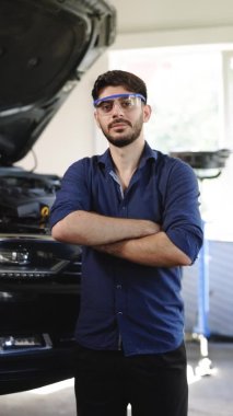 Vertical Format Video of Bearded Handsome Car Mechanic is Posing in a Car Service. He Wears a Jeans Shirt and Safety Glasses. His Arms are Crossed. Specialist Manager Looks at Camera