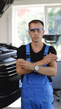 Vertical Video Portrait of Handsome Professional Car Mechanic Crossed Arms is Working on a Vehicle in a Service. Repairman Specialist is Wearing Safety Glasses Looks at a Camera.