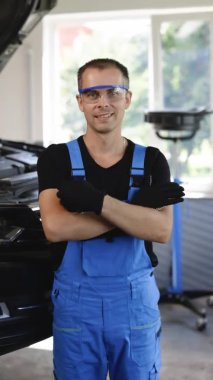 Vertical Video Portrait of Handsome Repairman Specialist Car Mechanic Wearing Safety Glasses Crossed Hands Looks at a Camera and Smile