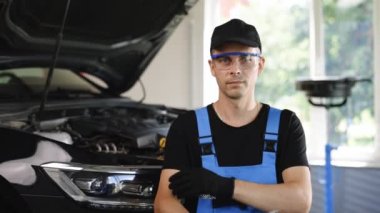 Portrait of handsome automotive mechanic man in blue uniform and cap standing in garage. Vehicle service manager worker work in mechanics workshop, looking at camera after check the engine.