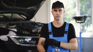 Portrait of handsome automotive mechanic man in blue uniform and black cap crossed arms standing in garage. Vehicle service manager worker work in mechanics workshop, looking at camera.