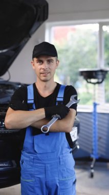 Vertical portrait shot of handsome caucasian young man in blue uniform smiling to camera and holding wrench. Male mechanic demonstrating spanner. Workday. Repair concept.