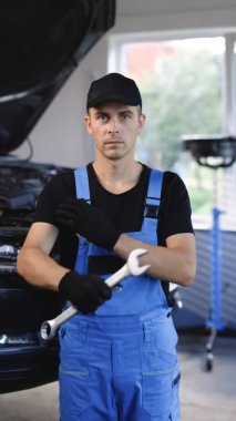 Vertical portrait shot of handsome caucasian man in blue uniform and black cap holding wrench and look to camera. Male mechanic demonstrating spanner. Workday. Repair concept.