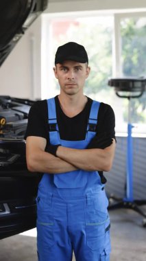 Vertical video portrait of handsome automotive mechanic man in blue uniform and black cap crossed arms standing in garage. Vehicle service manager worker work in mechanics workshop, looking at camera.