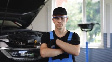 Portrait shot of caucasian man in blue uniform and hat standing in big auto garage, looking to camera and crossing hands. Pretty male mechanic in maintaining service.
