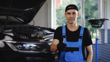 Portrait of car mechanic in a car workshop shows thumbs up, in the background of service. Positive auto service worker smiling to camera and showing thumb up gesture, approving car repair workshop