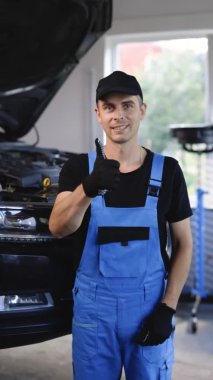 Vertical video of car mechanic in car workshop shows thumbs up. Positive auto service worker smiling to camera and showing thumb up gesture, approving car repair workshop. Workday. Repair concept.