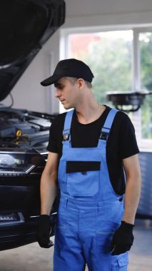 Vertical video young car mechanic working by showing thumb up by looking at camera at garage. Concept of approval, car repair service and professional worker.