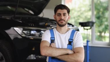 Portrait of bearded car mechanic crosses hands in a car workshop in blue uniform with equipment looking into camera. Male car mechanic at workplace in spacious repair shop.