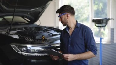 Portrait of happy cheerful Caucasian bearded man auto mechanic or manager smiling to camera and giving thumb up in garage. Young guy wears jeans shirt and safety glasses at cars service garage.