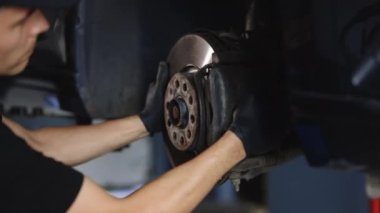 Mechanic at service station checks the brake and steering systems. Strong man checks for wear in running gear and steering components. Service operator in overalls. Car moves on lift.