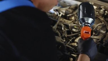 Caucasian Mechanic in Blue Overalls is Working on a Car in a Car Service. Repairman in Safety Glasses is Working on an Usual Car Maintenance. He Hangs Led Lamp. Modern Workshop.