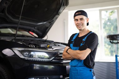 Portrait happy mechanic worker in blue uniform and cap from auto service center in front of the camera looking straight smiling and enjoying the time at his work place.