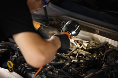 Repairman in Safety Glasses is Working on an Usual Car Maintenance. Mechanic is Working on a Car in a Car Service. He Hangs Led Lamp. Modern Workshop.