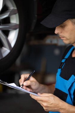 Car service employe inspect car. Mechanic inspects the car undercarriage way and makes a note on his inspection sheet. Automobile service, car mechanic. Modern workshop