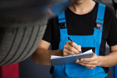 Car service employe inspect car. Mechanic inspects the car undercarriage way and makes a note on his inspection sheet. Automobile service, car mechanic. Modern workshop