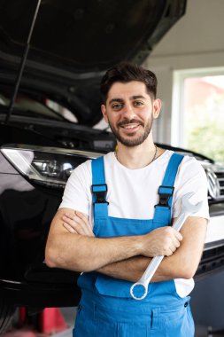 Caucasian bearded man in blue coveralls holds spanner, smiling and looking into camera. Male car mechanic in spacious repair shop.