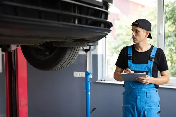 Automobile service, car mechanic. Car mechanic at a repair shop taking notes on his tablet. Car service employe inspect car. Modern clean workshop.