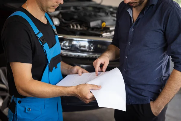 Specialist is showing info on tablet. Car service manager with a tablet, talking to mechanic man discussing car diagnostics and repairing. Mechanic talking to manager near vehicle in car service.