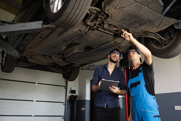 Auto Service. Car Service Employees Inspect the Bottom and Skid Plates of the Car. Manager Checks Data on a Notebook and Explains the Breakdown to a Mechanic. Modern Workshop.