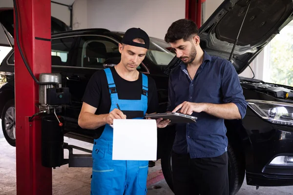 Car Service Employees Inspect the Bottom and Skid Plates of the Car. Manager Checks Data on a Tablet Computer and Explains the Breakdown to Mechanic. Specialist is Showing Info on Tablet