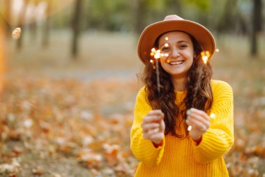 Ellerinde pırıl pırıl parlıyor. Doğada Noel havai fişekleri olan sarı kazaklı genç bir kadın. Sonbahar stili. 