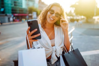 Telefonu kullanan, alışveriş torbalarını tutan, dışarıda duran, kıyafet ve hediye seçen genç bir kadın. İnternetten alışveriş konsepti. Tüketim ve Teknoloji.