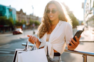 Telefonu kullanan, alışveriş torbalarını tutan, dışarıda duran, kıyafet ve hediye seçen genç bir kadın. İnternetten alışveriş konsepti. Tüketim ve Teknoloji.