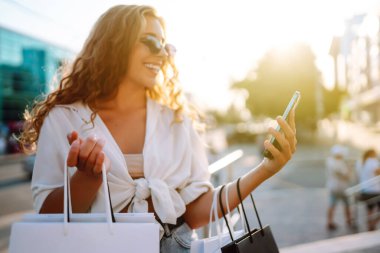 Telefonu kullanan, alışveriş torbalarını tutan, dışarıda duran, kıyafet ve hediye seçen genç bir kadın. İnternetten alışveriş konsepti. Tüketim ve Teknoloji.