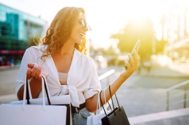 Telefonu kullanan, alışveriş torbalarını tutan, dışarıda duran, kıyafet ve hediye seçen genç bir kadın. İnternetten alışveriş konsepti. Tüketim ve Teknoloji.