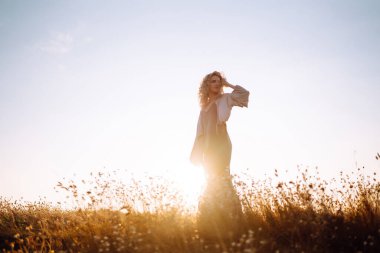 Kıvırcık kadın denizin üstünde bir kayanın kenarında poz veriyor, mavi gökyüzü ve deniz altyapısı ile. Sonbahar tarzı. Seyahat ve macera.