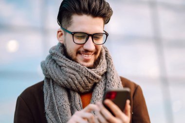 Hat bağlantısı. Elinde akıllı telefon olan paltolu genç bir adam sokakta yürüyor. Şehir geçmişinde modern saç stili olan sakallı genç adam cep telefonuyla yazı yazıyor.. 