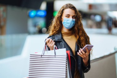 Alışveriş torbası tutarken cep telefonu kullanan koruyucu maskeli genç bir kadın. Ticaret, alıcılar, kara cuma, indirimler, satış konsepti. Coronavirüs salgınında alışveriş..