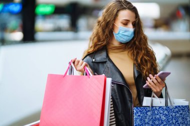 Alışveriş torbası tutarken cep telefonu kullanan koruyucu maskeli genç bir kadın. Ticaret, alıcılar, kara cuma, indirimler, satış konsepti. Coronavirüs salgınında alışveriş..