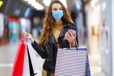 Alışveriş torbası tutarken cep telefonu kullanan koruyucu maskeli genç bir kadın. Ticaret, alıcılar, kara cuma, indirimler, satış konsepti. Coronavirüs salgınında alışveriş..