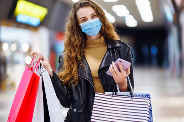 百貨店でショッピングバッグを保持しながら携帯電話を使用して保護マスクの若い女性 バイヤー 黒金曜日 販売コンセプト コロナウイルス流行のショッピング — ストック写真