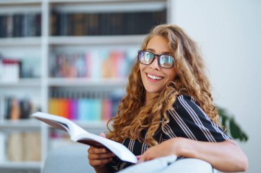 Kitapla oturan genç bir kadın, kütüphanede laptok oturup öğrenme notları alıyor. Eğitim kavramı, kütüphane, öğrenciler. 