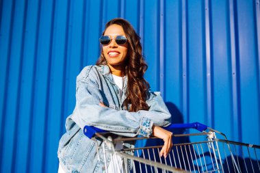 Alışveriş arabalı genç bir kadın renk duvarının yanında eğleniyor. Gülümseyen Afrikalı kadın alışverişe çıkıyor. Kara Cuma. Tüketim, satış, indirimler, yaşam tarzı konsepti.