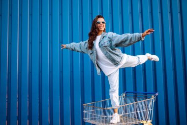 Alışveriş arabalı genç bir kadın renk duvarının yanında eğleniyor. Gülümseyen Afrikalı kadın alışverişe çıkıyor. Kara Cuma. Tüketim, satış, indirimler, yaşam tarzı konsepti.