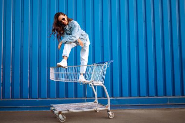 Alışveriş arabalı genç bir kadın renk duvarının yanında eğleniyor. Gülümseyen Afrikalı kadın alışverişe çıkıyor. Kara Cuma. Tüketim, satış, indirimler, yaşam tarzı konsepti.