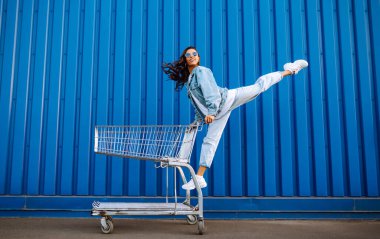 Alışveriş arabalı genç bir kadın renk duvarının yanında eğleniyor. Gülümseyen Afrikalı kadın alışverişe çıkıyor. Kara Cuma. Tüketim, satış, indirimler, yaşam tarzı konsepti.