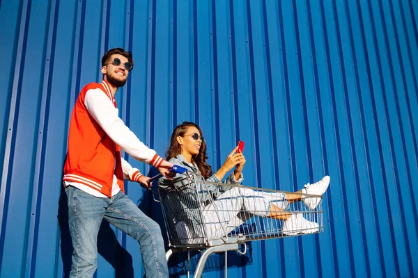 Pozitif erkek ve kadın eğleniyor ve alışveriş arabasına biniyor. İki genç arkadaş alışverişe gidiyor. Kara Cuma. Tüketim, satış, indirimler, yaşam tarzı konsepti.
